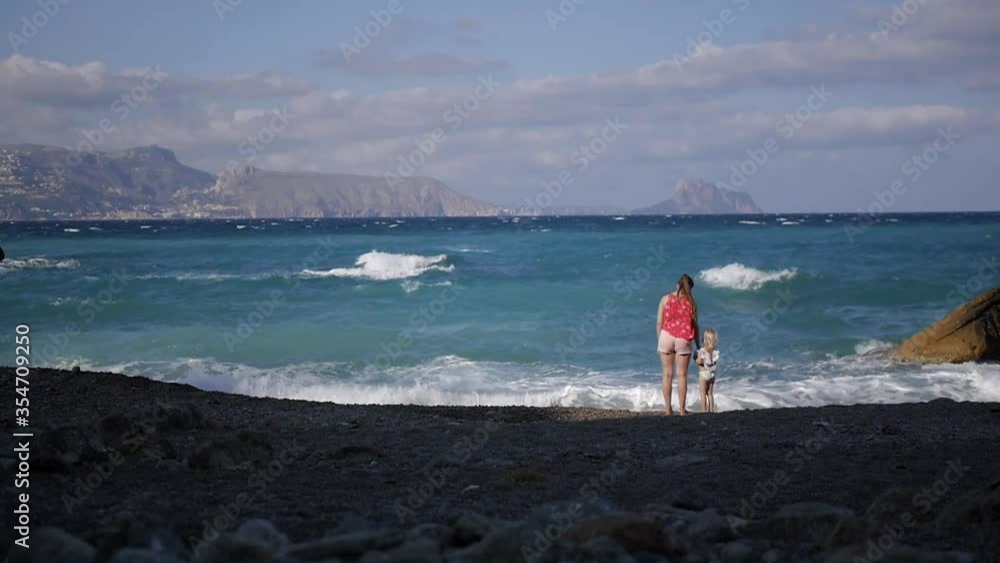 Young woman in protective mask with child on the beach. Little blond girl hold mother's hand standing in water. Walking by the sea during pandemic. Wide shot. Young woman in protective mask with child on the beach. Little blond girl hold mother's hand standing in water. Walking by the sea during pandemic. Wide shot.