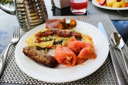 This is breakfast at a five star hotel. Salmon, sausage, egg omelet are on a plate on the table. Fork, knife and spoon, carafe and a glass with tomato juice, Aruba, August 2014