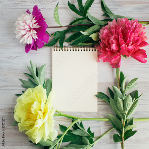 A blank notebook surrounded by colorful flowers of peonies, a light wooden background, To-do list, plans, dreams. Top view, Vintage toned. Copy space for text