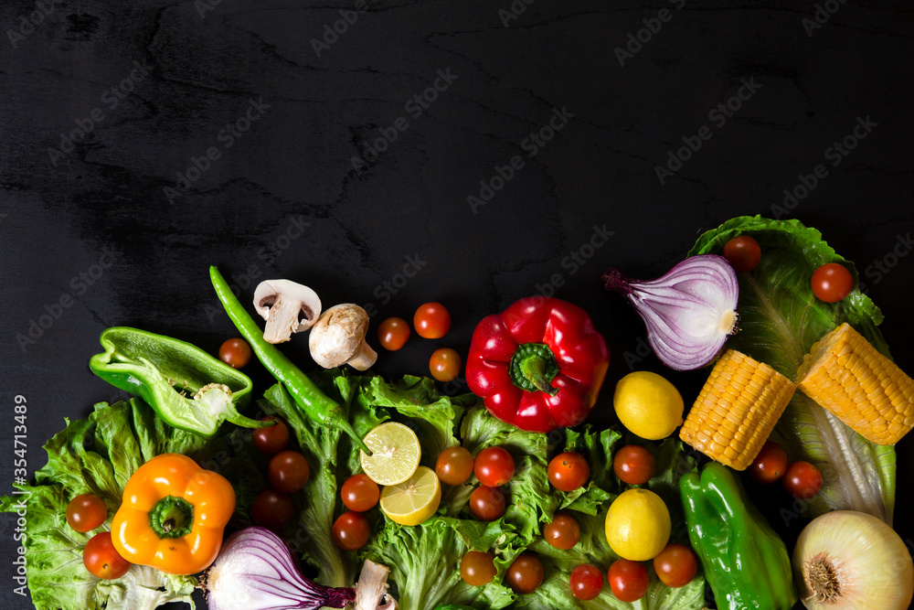 Various fresh vegetables arranged on black surface. Top view of pepper, corn, cherry tomatoes, mushrooms, lemon, onion and lettuce arranged on black table.
