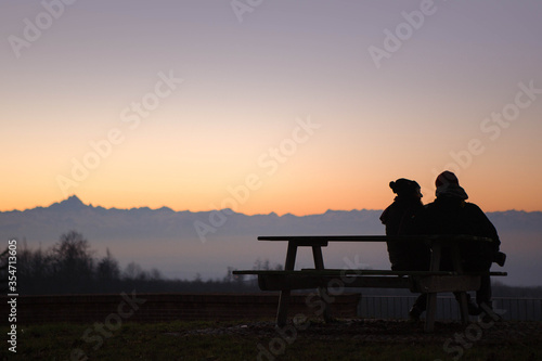 silhouettes of a Couple sitting in a bench enjoying the wonderful color of the sky at dusk, in the background the top of the pink mountain, Italy.