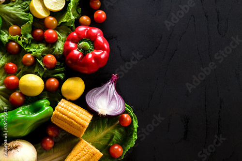 Vegetables arranged on black table with empty space on right. Black surface with green pepper, corn, cherry tomatoes, lemons, onions and lettuce.