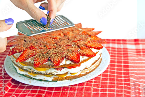 Woman is grating chocolate on home made cake with strawberry, peach, whipped cream - delicious bakery for celebration