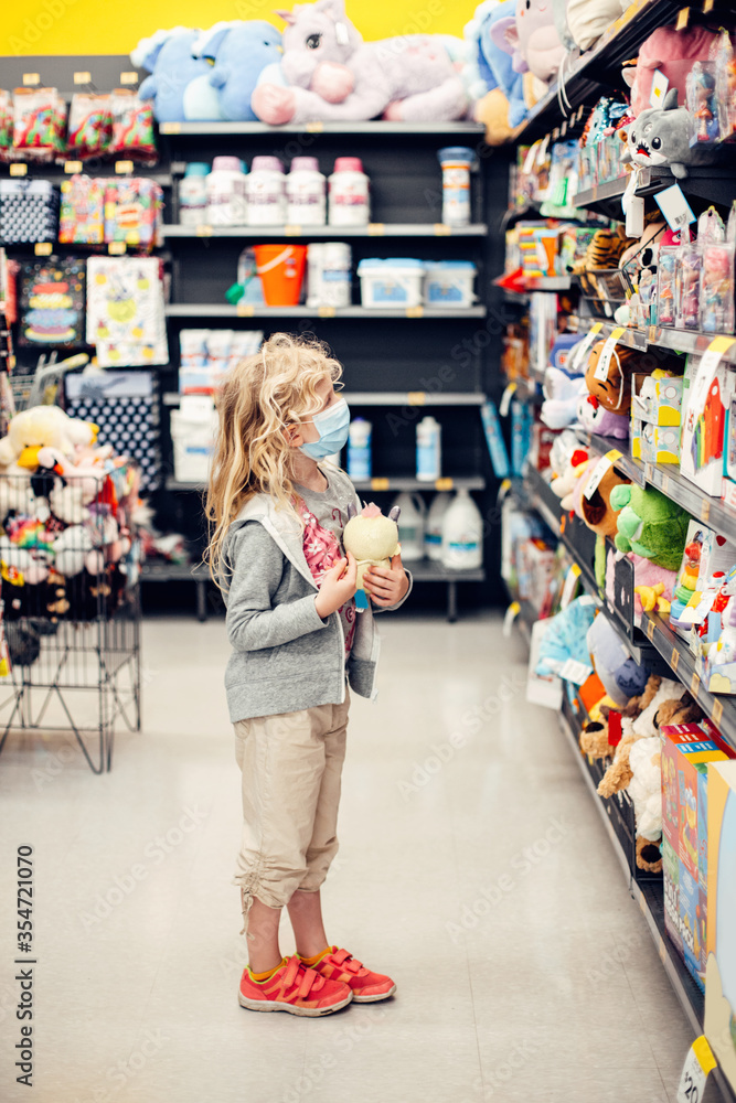 A new normal. Caucasian blonde girl in sanitary face mask shopping at toy store. Child wearing protective mask against coronavirus. Safety, health protection during covid-19 quarantine. A new normal. Caucasian blonde girl in sanitary face mask shopping at toy store. Child wearing protective mask against coronavirus. Safety, health protection during covid-19 quarantine.