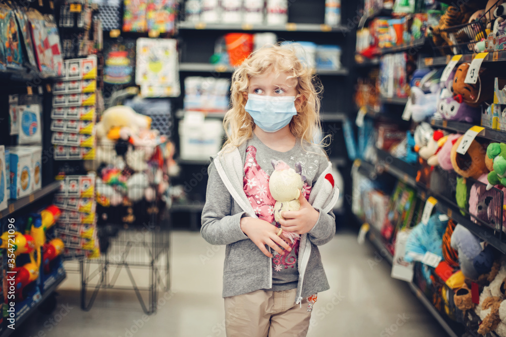 Obraz premium A new normal. Caucasian blonde girl in sanitary face mask shopping at toy store. Child wearing protective mask against coronavirus. Safety, health protection during covid-19 quarantine.