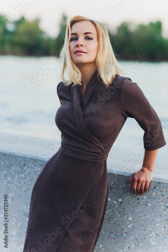 Outdoor portrait of young beautiful caucasuan woman with long straight hair in short black dress, beige shoes holding brown leather handbag walking city street. photo