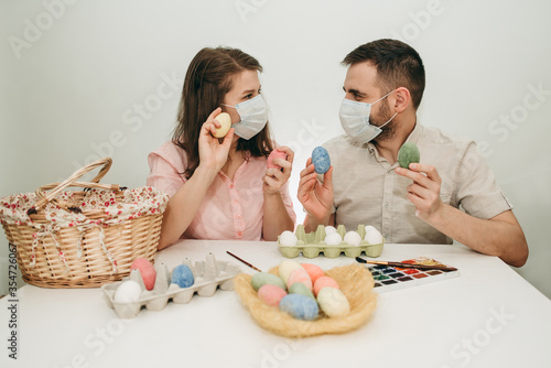 Young white European couple celebrating paint Easter quarantine at home in medical surgical masks. Basket, eggs, holiday, coronavirus, illness, stay home, infection, flue, surgical bandage.