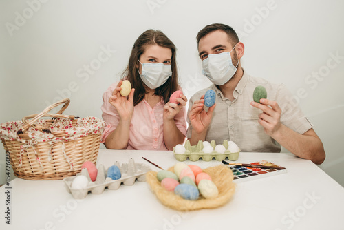 Young white European couple celebrating paint Easter quarantine at home in medical surgical masks. Basket, eggs, holiday, coronavirus, illness, stay home, infection, flue, surgical bandage.
