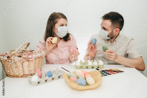 Young white European couple celebrating paint Easter quarantine at home in medical surgical masks. Basket, eggs, holiday, coronavirus, illness, stay home, infection, flue, surgical bandage.