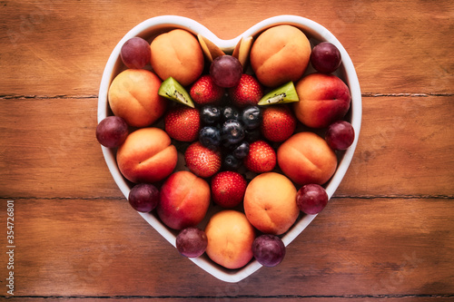 Close-up view from above of a white heart-shaped plate full of red grapes  apricots  strawberry and blueberries. Wooden background