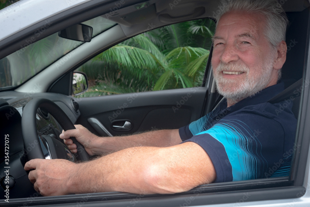 Obraz premium Attractive senior man smiles after parking the car. Caucasian senior people with gray hair and beard. Green plants outside the car window