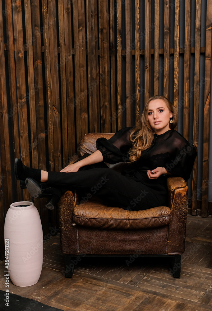 Elegant blonde lady posing in black blouse with shoes heels. Fashionable woman sitting on big leather chair. Wooden wall on the background. Elegant blonde lady posing in black blouse with shoes heels. Fashionable woman sitting on big leather chair. Wooden wall on the background.