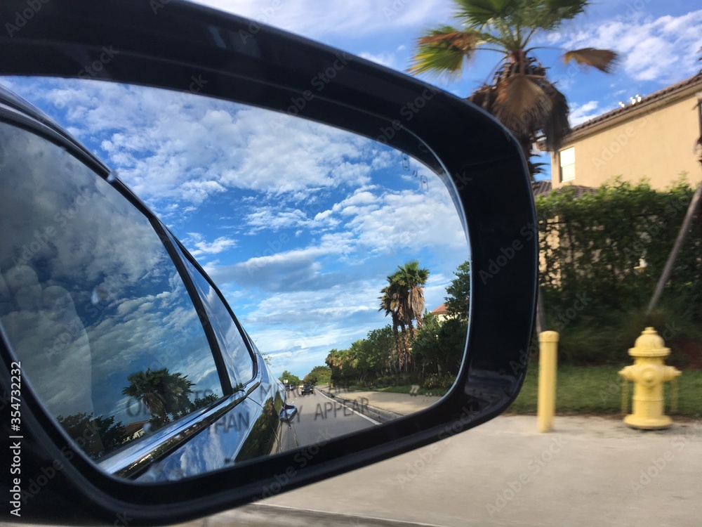 Traveling automobile car closeup of side view mirror driving past green Florida palm tree street landscape and cloudy blue skies on highway