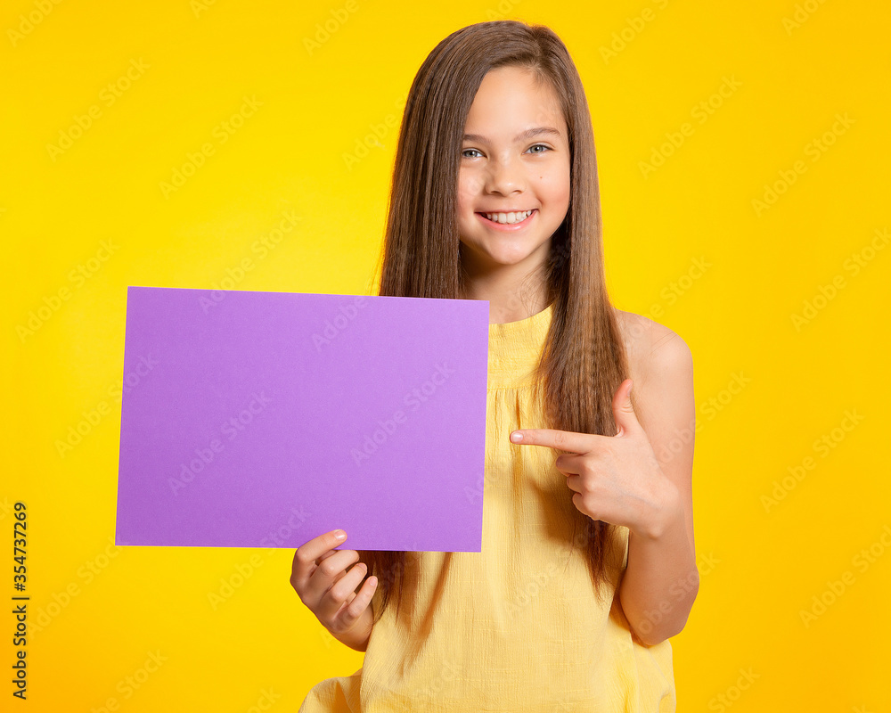 A beautiful European brunette teen with blue eyes holds a purple sheet of paper on a yellow background, smiles and points at it. Space for text, banner, surface for labels and ads. A beautiful European brunette teen with blue eyes holds a purple sheet of paper on a yellow background, smiles and points at it. Space for text, banner, surface for labels and ads.