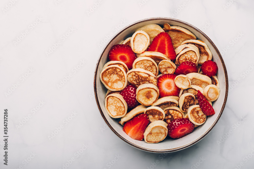 Trendy home breakfast with tiny cereal pancakes with piece of butter and fresh strawberries in the bowl. View from above. Trendy home breakfast with tiny cereal pancakes with piece of butter and fresh strawberries in the bowl. View from above.