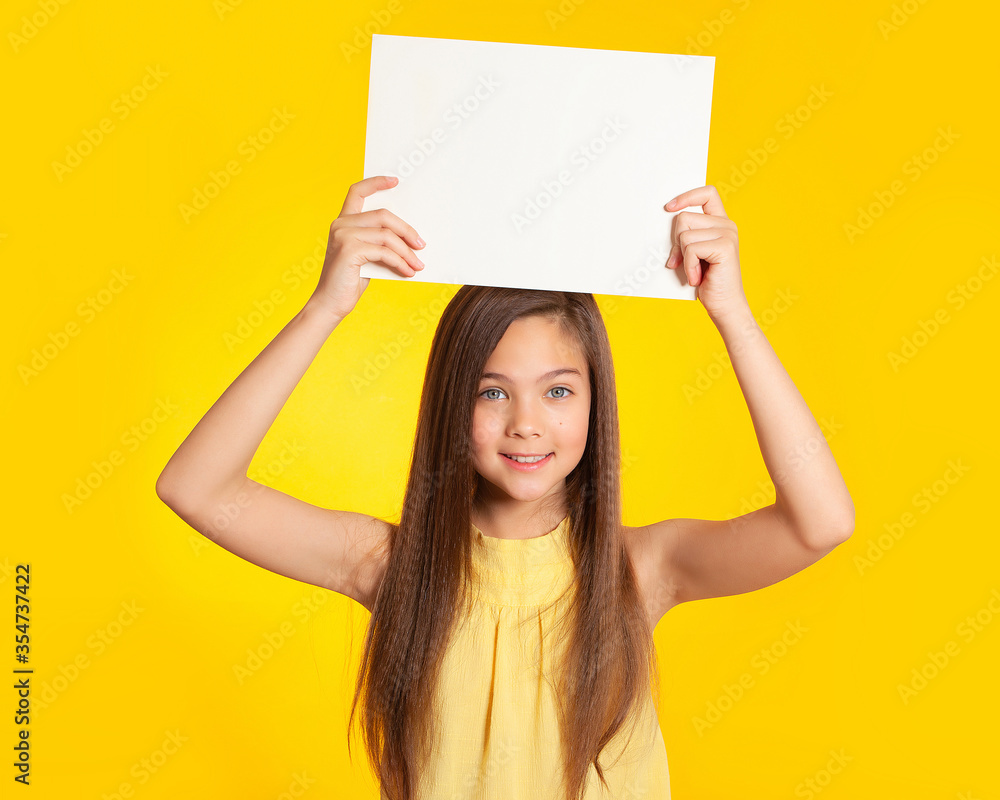 Beautiful European brunette teenager girl with blue eyes holds a white sheet of paper over her head on a yellow background and smiles. Place for text, banner, surface for inscriptions and advertising. Beautiful European brunette teenager girl with blue eyes holds a white sheet of paper over her head on a yellow background and smiles. Place for text, banner, surface for inscriptions and advertising.