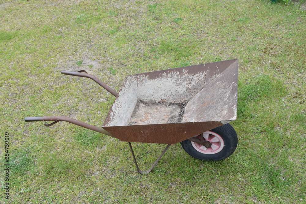 An empty wheelbarrow standing on freshly cut grass, burnt in places by the sun. A rusty, one-wheelbarrow used for gardening. An empty wheelbarrow standing on freshly cut grass, burnt in places by the sun. A rusty, one-wheelbarrow used for gardening.