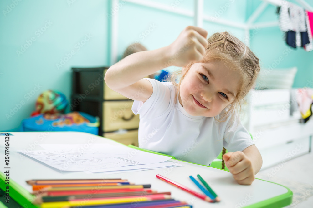 Little girl draws while sitting at a table in a room against the background of the wall Little girl draws while sitting at a table in a room against the background of the wall