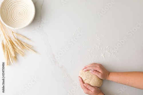 Top view of female hand molding bread dough in a marble table with an empty bread bowl and wheat with space for text