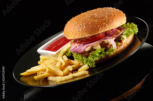 Big fresh burger with sauce and fries on black plate isolated on black background.  American classic burger menu.