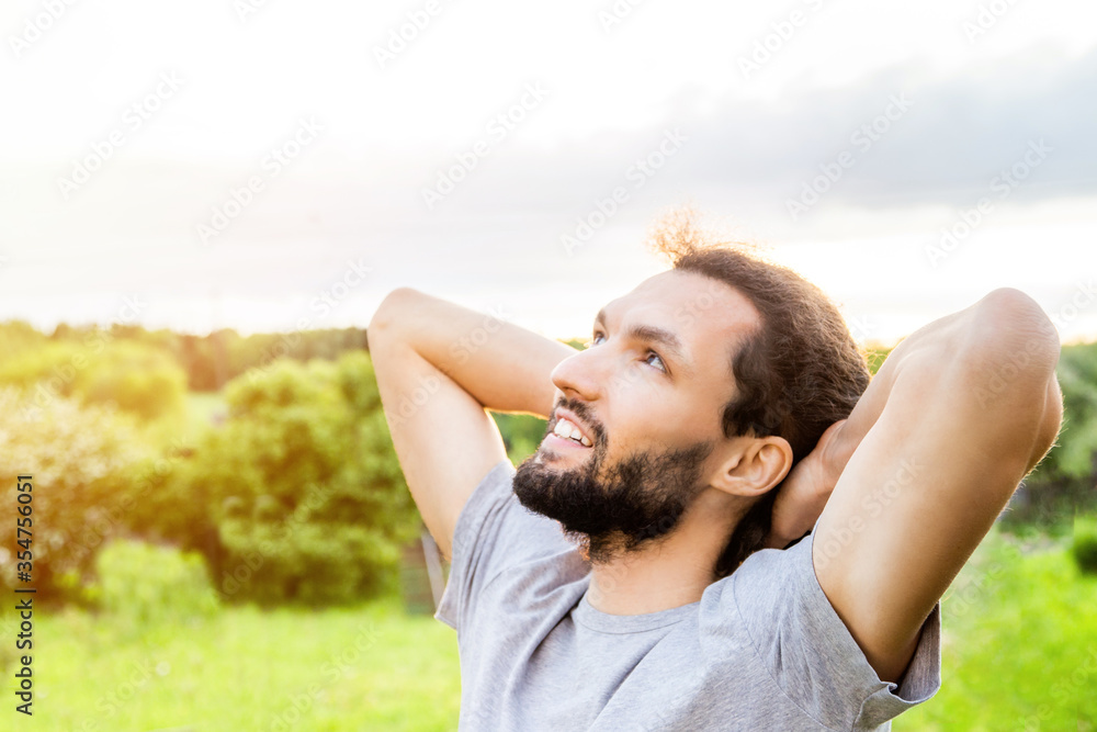 Happy young breaded man relaxing outdoors with hands behind head, nature and vegetation on background. Enjoy the summer air and relax in the countryside.