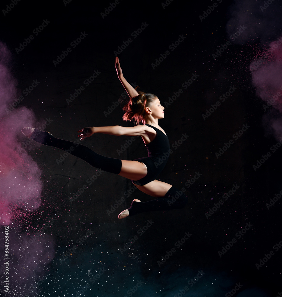 Young girl gymnast in black sport body and special uppers jumping in gymnastic pose over dark background