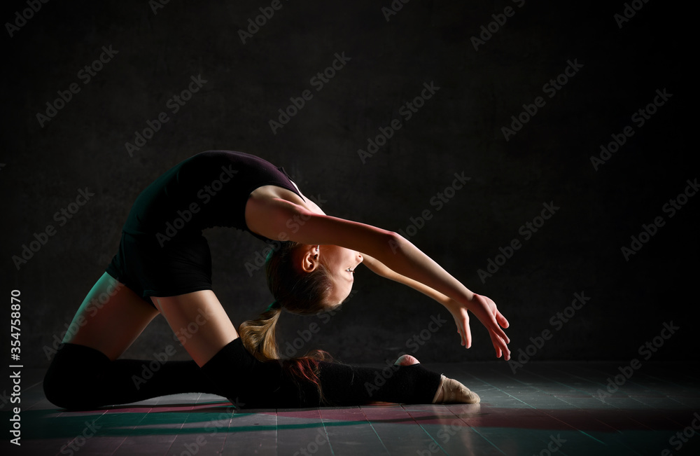 Young girl gymnast in black sport body and special footwear making gymnastic pose over dark background
