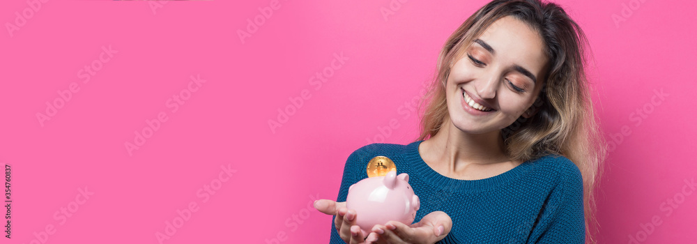Girl puts in the piggy bank physical Bitcoin.Young girl over pink background holding piggy bank and rejoices gesturing. The concept of reliability of cash investments and insurance. Girl puts in the piggy bank physical Bitcoin.Young girl over pink background holding piggy bank and rejoices gesturing. The concept of reliability of cash investments and insurance.