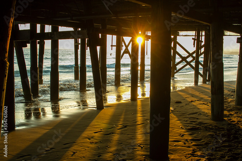 Sunset from under a pier with surf sand waves and surfers out in the water