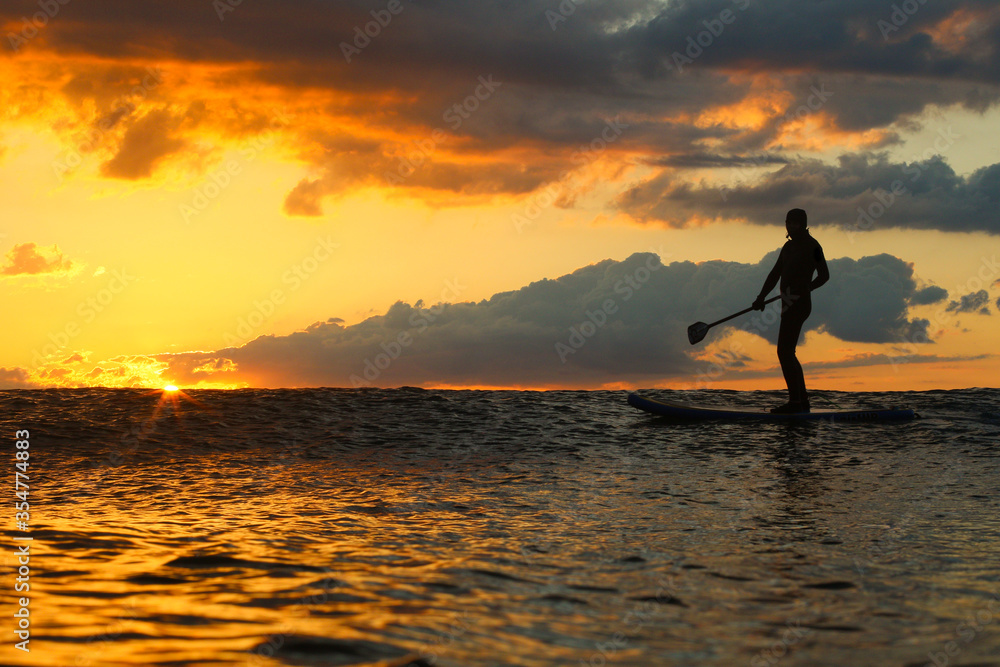 Stand Up Paddle Boarding In Japan at Sunrise and Sunset a solo rider keeping fit & healthy on the Pacific Ocean in a black wetsuit, also catching some large waves, The ocean is blue with a nice sky.