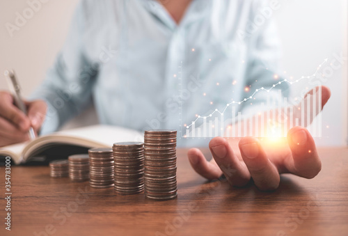 Businessman holding virtual graph and writing saving dividend or profit to notebook with coins stacking . Business investment and saving profit concept.