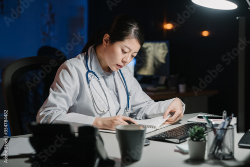asian japanese female doctor writing on notepad in medical office at late night beautiful concentrated girl clinic worker sitting at desk reading notebook in dark workplace in hospital at midnight.