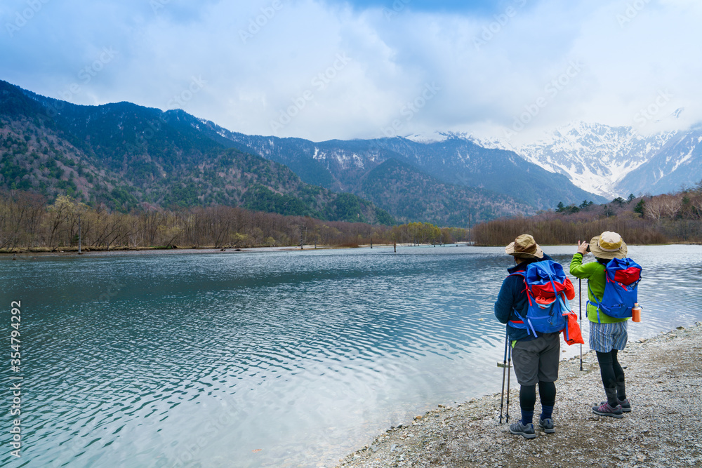 Fototapeta premium Hikers wearing colourful attire stop and photograph Lake Taisho and the distant snowy mountain peaks of Chubu Sangaku National Park in Kamikochi, Nagano Prefecture, Japan. Selective focus.