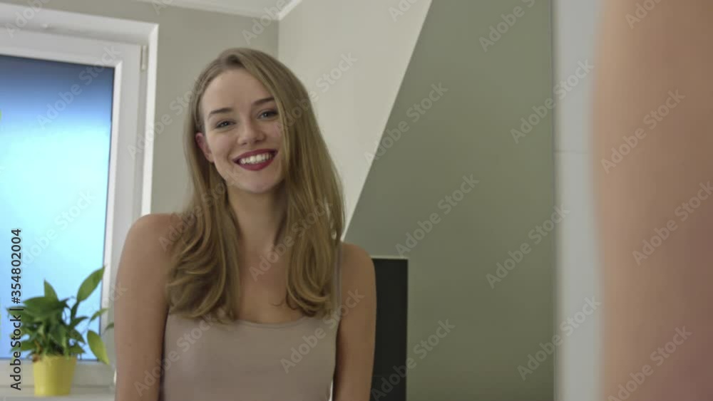 Happy beautiful woman smiling while looking at the mirror. Reflection of blonde girl as she smiles wearing casual inside a room. Indoor shot of attractive lady Happy beautiful woman smiling while looking at the mirror. Reflection of blonde girl as she smiles wearing casual inside a room. Indoor shot of attractive lady