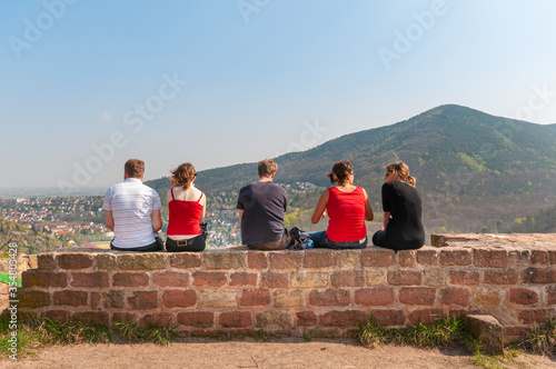 Germany. Four young friends, male and female, in the sun, sitting on a stone wall facing the landscape in front of them, after a nice walk during the summer. Boys and Grils seen from seen from behind. photo