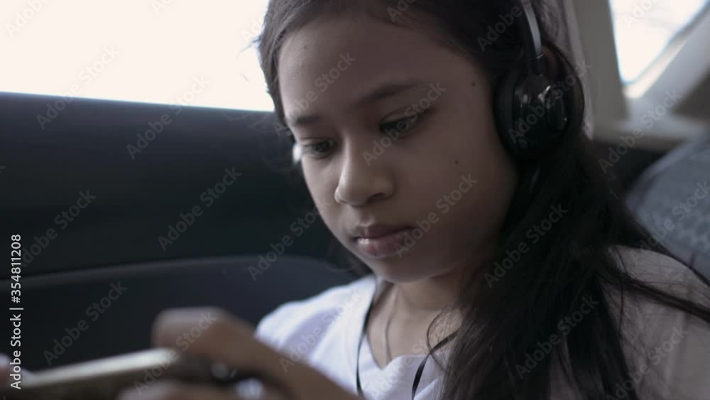Cute asian young girl wearing headphones and using mobile phones during traveling in a car. Serious female teen gamer playing online games while sitting in the back seat of a car. Cute asian young girl wearing headphones and using mobile phones during traveling in a car. Serious female teen gamer playing online games while sitting in the back seat of a car.