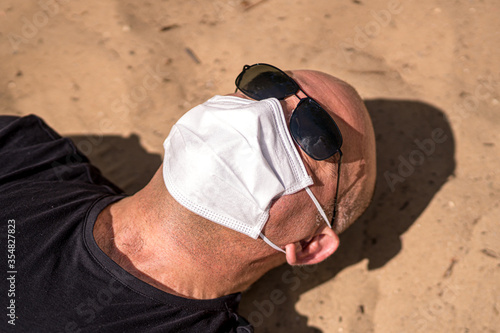 Summer holiday during the lockdown or the coronavirus disease outbreak. Man wearing sunglasses and medical protective mask sunbathing on a sandy beach