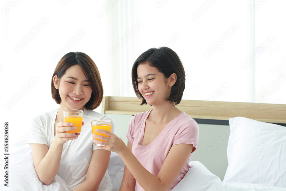 Asian young sisters lovely couple on white bed and smiling with hand holding Orange juice glass in bedroom. Holiday and healthy concept Asian young sisters lovely couple on white bed and smiling with hand holding Orange juice glass in bedroom. Holiday and healthy concept