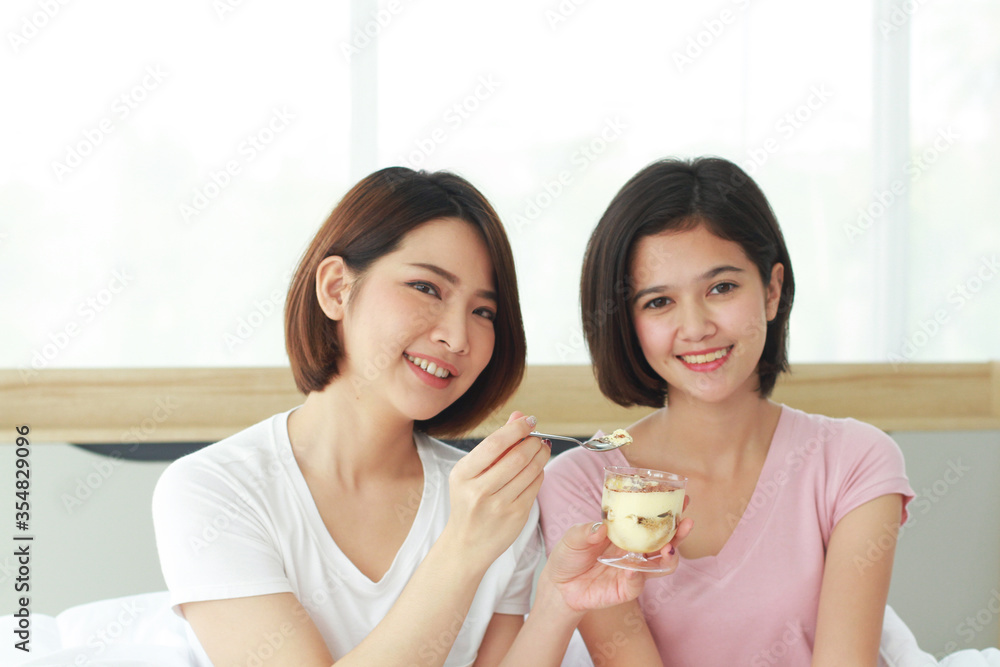 Asian young sisters lovely couple on white bed hand holding cup of cake and eating Bakery together in bedroom. Homosexual women or Lesbian in love. LGBT Asian and Holiday concept. Asian young sisters lovely couple on white bed hand holding cup of cake and eating Bakery together in bedroom. Homosexual women or Lesbian in love. LGBT Asian and Holiday concept.