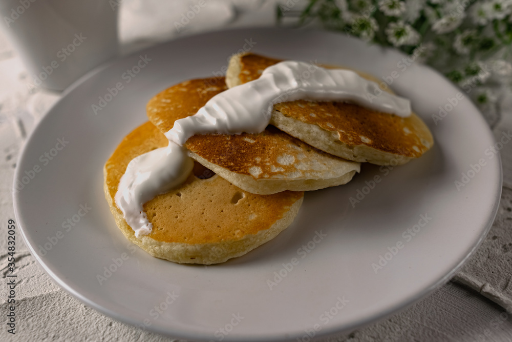 Crumpets and pancakes with sour cream on a white plate with white sauce on top. Next to a sprig of fresh light flowers and compote. Interesting textured background of white stone Crumpets and pancakes with sour cream on a white plate with white sauce on top. Next to a sprig of fresh light flowers and compote. Interesting textured background of white stone