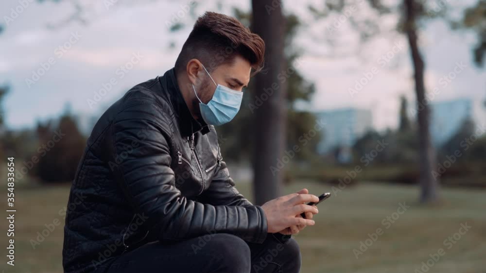 A handsome man with a surgical mask sits on the bench in the park. Make a bank transfer using your mobile phone. Covid-19. 4k A handsome man with a surgical mask sits on the bench in the park. Make a bank transfer using your mobile phone. Covid-19. 4k