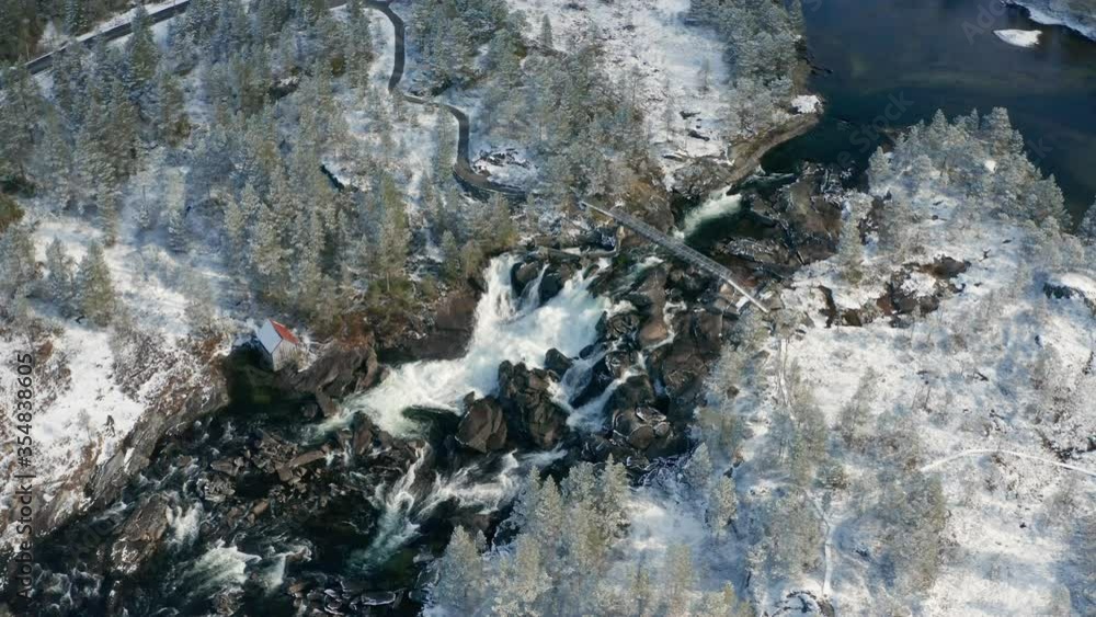 Aerial view of the Likholefossen waterfall on the Gaula river in Norway. A turbulent mass of water rushing down in the narrow rocky watercourse. A narrow steel bridge hangs above the waterfall. Aerial view of the Likholefossen waterfall on the Gaula river in Norway. A turbulent mass of water rushing down in the narrow rocky watercourse. A narrow steel bridge hangs above the waterfall.