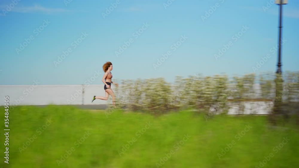 Side view tracking wide shot of determined young woman with curly red hair running fast on embankment during cardio workout on summer day Side view tracking wide shot of determined young woman with curly red hair running fast on embankment during cardio workout on summer day