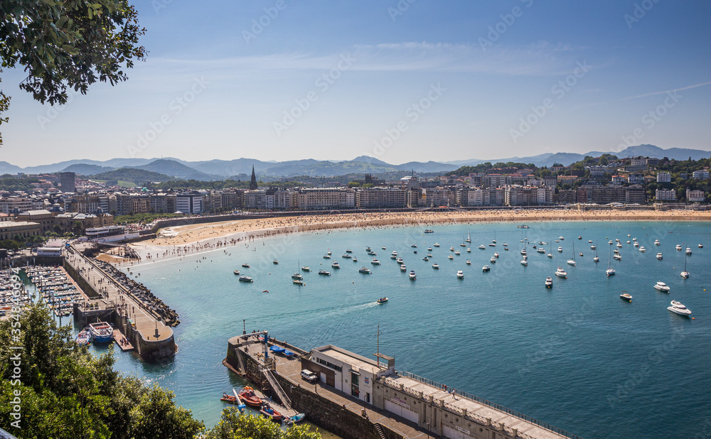 Panoramic view of a bay with a small port and a beach with people swimming, with some boats in the sea on a quiet summer day Panoramic view of a bay with a small port and a beach with people swimming, with some boats in the sea on a quiet summer day