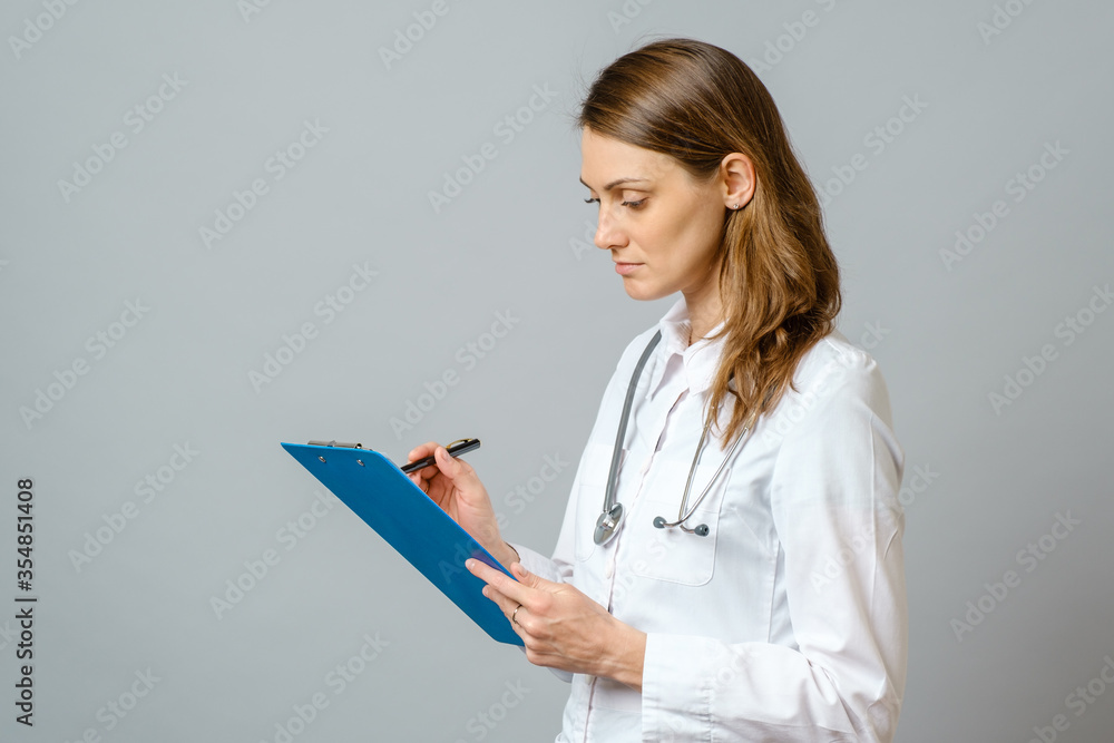 Young woman physician with stethoscope prescribing treatment to patient. Female doctor with pen writing recipe on clipboard. Isolated on gray Young woman physician with stethoscope prescribing treatment to patient. Female doctor with pen writing recipe on clipboard. Isolated on gray