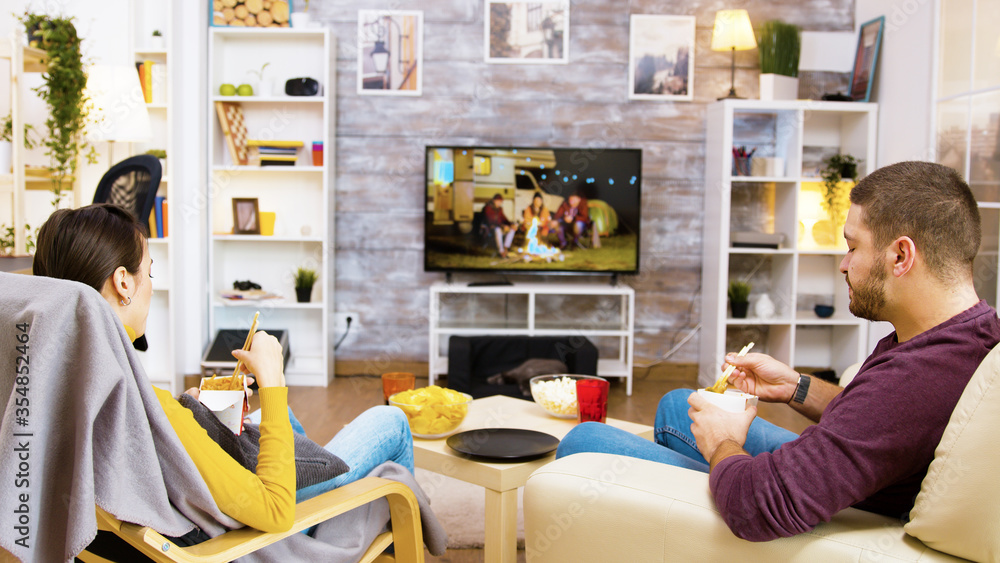 Back view of young couple sitting on comfortable chairs eating noodles in front of tv using chopsticks. Cat sleeping on her cozy bed. Back view of young couple sitting on comfortable chairs eating noodles in front of tv using chopsticks. Cat sleeping on her cozy bed.