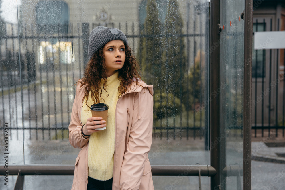 Fototapeta premium Sad young curly woman with cup of coffee stay at the bus stop in rainy day 