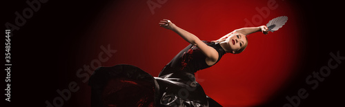 panoramic shot of young flamenco dancer in dress holding fan while dancing on red