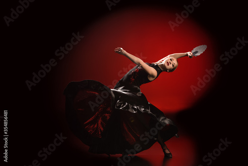 young flamenco dancer in dress holding fan while dancing on red and black photo