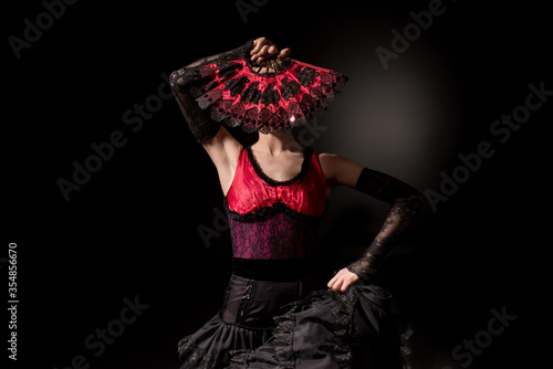 young flamenco dancer in dress covering face with fan on black